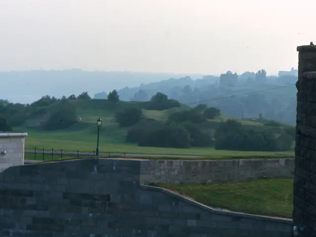 In this image there are buildings, wall, lamp, railing, trees, grass, mountains and the sky.