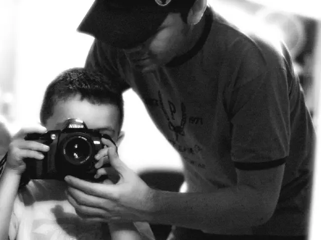 In this image I can see a boy is holding a camera, here I can see a person is standing.